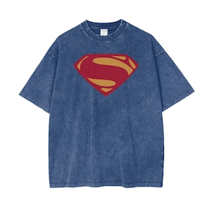 Könnte beinhalten: Ein kurzärmeliges T-Shirt in blauer Acid-Wash-Optik mit einem großen, mittig platzierten Superman-Logo. Das ikonische Emblem besteht aus einer dunkelroten Rautenform mit einer goldfarbenen S-förmigen Grafik in der Mitte, die auf der Brust aufgedruckt ist.
