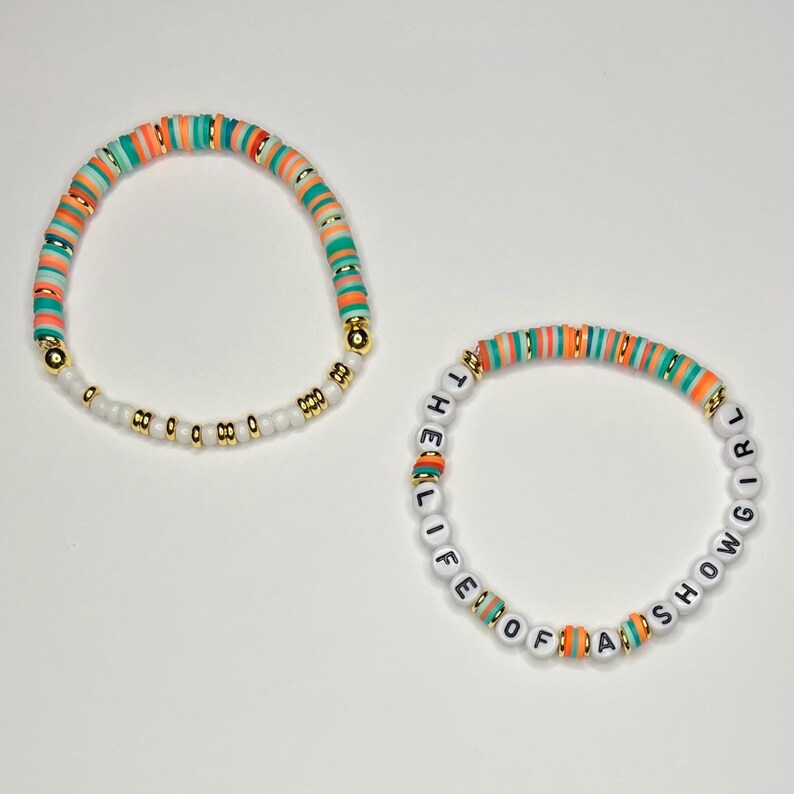 May include: Two beaded bracelets. One bracelet features orange, teal, and gold beads. The other bracelet has white beads with the text "THE LIFE OF A SHOW GIRL" and orange, teal, and gold beads.