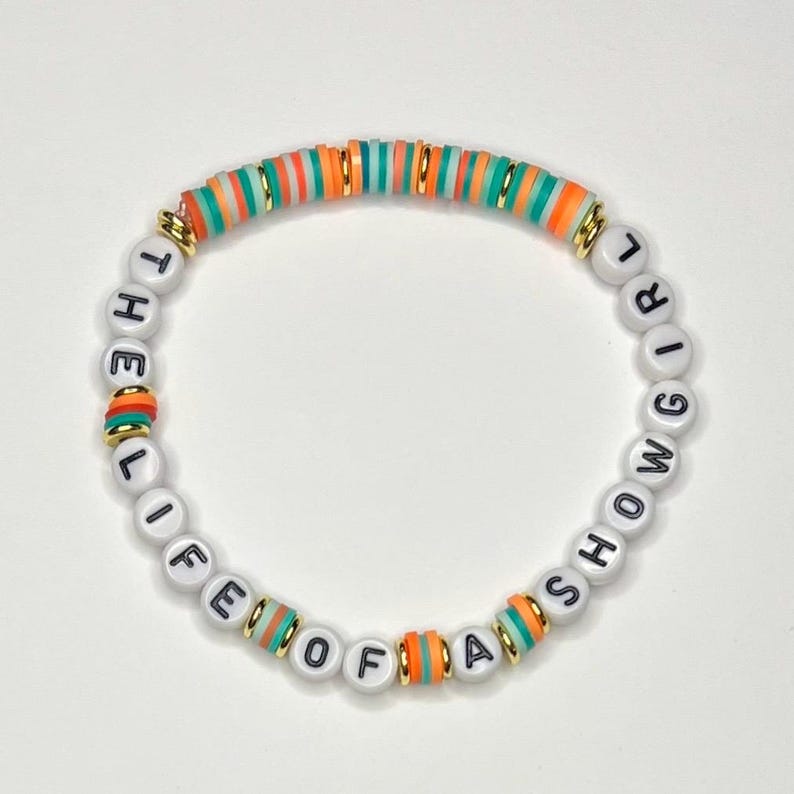 May include: A beaded bracelet with the text "THE LIFE OF A SHOWGIRL" in black letters on white beads. The bracelet features colorful orange, green, and gold accents.