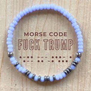 May include: A light purple beaded bracelet with the text "MORSE CODE FUCK TRUMP". The bracelet has silver-coloured metal accents and is displayed on a wooden surface. The Morse code is also shown.