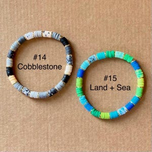 May include: Two beaded bracelets. One is labeled "#14 Cobblestone" with gray, black, white, and gold beads. The other is labeled "#15 Land + Sea" with blue, green, and gold beads. Both bracelets are circular.