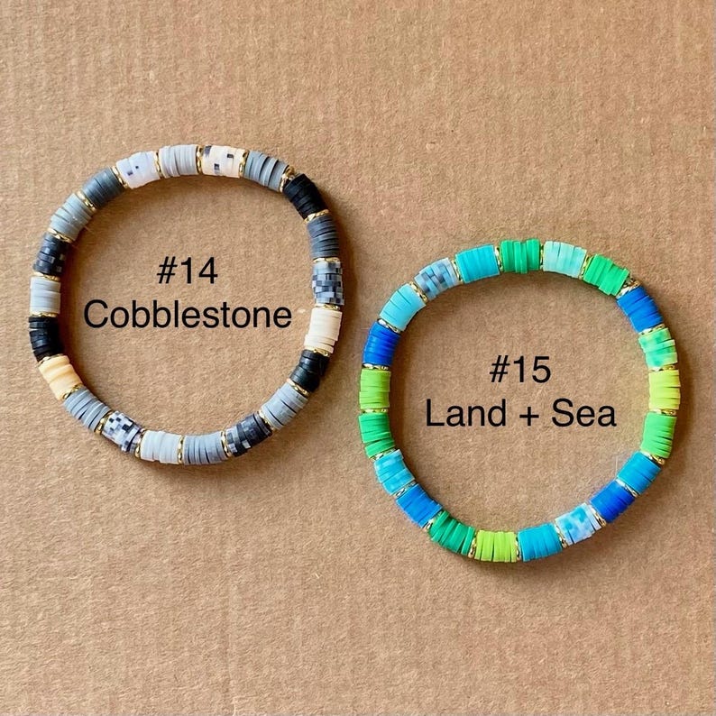 May include: Two beaded bracelets. One is labeled "#14 Cobblestone" with gray, black, white, and gold beads. The other is labeled "#15 Land + Sea" with blue, green, and gold beads. Both bracelets are circular.