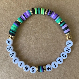 May include: A colorful beaded bracelet with black, green, and purple stripes. The bracelet has white beads with the words "Feeling Witchy" spelled out.