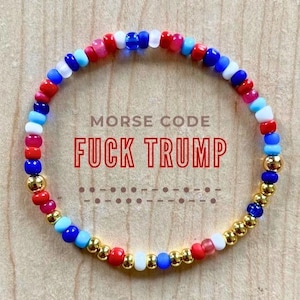 Peut inclure: Un bracelet de perles avec des perles rouges, blanches, bleues et dorées. Le texte "MORSE CODE FUCK TRUMP" est affiché en rouge, avec une représentation en code Morse en dessous. Le bracelet est sur une surface en bois.