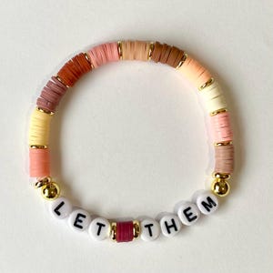 May include: A bracelet with a gradient of pink, orange, and brown beads. The bracelet has gold accents and the words "LET THEM" spelled out in white beads.