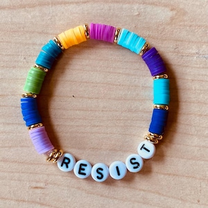 May include: A colorful bracelet with rainbow-colored beads and gold accents. The bracelet spells out the word "RESIST" in white letters with black outlines.