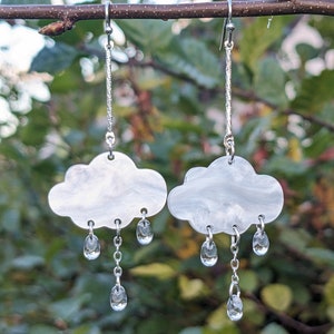 Può includere: Un paio di orecchini pendenti argentati con un ciondolo a forma di nuvola bianca, una catena argentata e accenti di gocce di pioggia di cristallo trasparente.