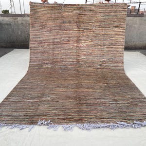 May include: A large, rectangular rug with a horizontal striped pattern in shades of brown, beige, and hints of blue and red. The rug has a fringe along the bottom edge. The rug is displayed on a white surface.