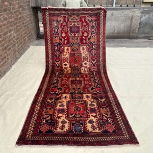 May include: A rectangular, hand-knotted rug with a central design in red, beige, and navy blue. The rug features intricate geometric patterns and a detailed border. The rug is displayed outdoors, showcasing its full length.