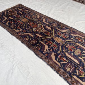 May include: A navy blue runner rug with a detailed pattern in beige, red, and green. The rug's central design features geometric and floral motifs, framed by a patterned border. The rug is displayed on a white surface.