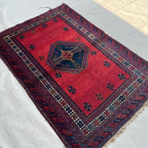 May include: A rectangular, hand-woven rug with a red field and a dark blue central medallion. The rug features a detailed border with geometric patterns in shades of blue, brown, and white. The rug's dimensions are not visible.