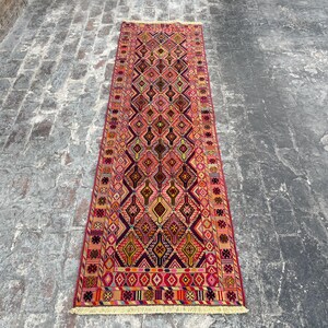 May include: A long, narrow rug with a geometric pattern in shades of red, pink, yellow, and blue. The rug has a fringed border.