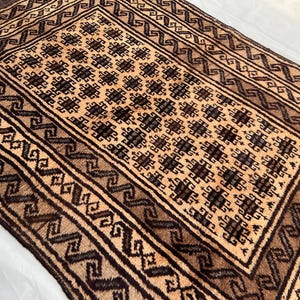 May include: A brown and beige rug with a geometric pattern. The rug has a central square with a repeating pattern of black and brown squares. The border of the rug is a repeating pattern of brown and beige.