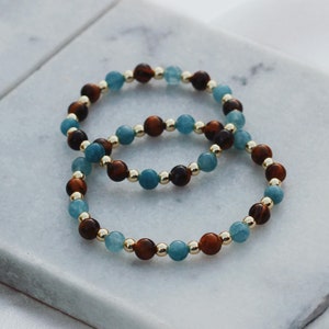 May include: Two beaded bracelets, one blue and brown, the other blue and gold, on a white marble surface.