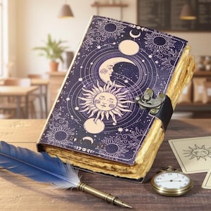 May include: A dark blue journal with celestial designs, including a sun and moon, sits open on a wooden table. A blue feather quill, pocket watch, and cards with sun and moon symbols are nearby. The journal has a leather strap closure.