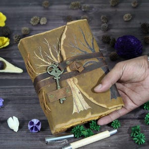 May include: A handmade leather journal with a vintage key closure. The journal is brown with a tree branch design and a leather strap. The key is antique brass. The journal is held by a person's hand.