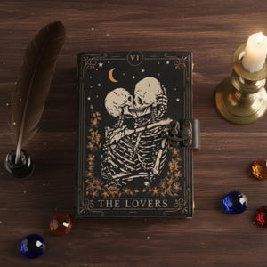 Könnte beinhalten: Ein schwarzes Notizbuch mit Metallverschluss, das ein Tarotkarten-Design von zwei sich umarmenden Skeletten zeigt, mit den Worten "THE LOVERS" unten. Eine brennende Kerze in einem Messinghalter, eine Feder und dekorative Edelsteine sind ebenfalls vorhanden.