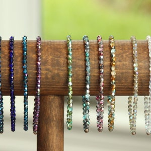 Può includere: Esposizione di braccialetti di perline in vari colori, tra cui rosso, blu, verde e oro, disposti su un supporto di legno. I braccialetti sono fatti di piccole perline sfaccettate, ognuna di un colore diverso. Il supporto è in legno scuro.
