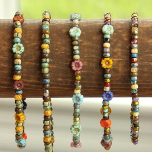 Puede incluir: Seis pulseras de cuentas coloridas con dijes en forma de flor. Las pulseras están hechas con cuentas pequeñas multicolores y presentan una variedad de dijes en forma de flor en diferentes colores, incluyendo turquesa, morado, naranja y amarillo.