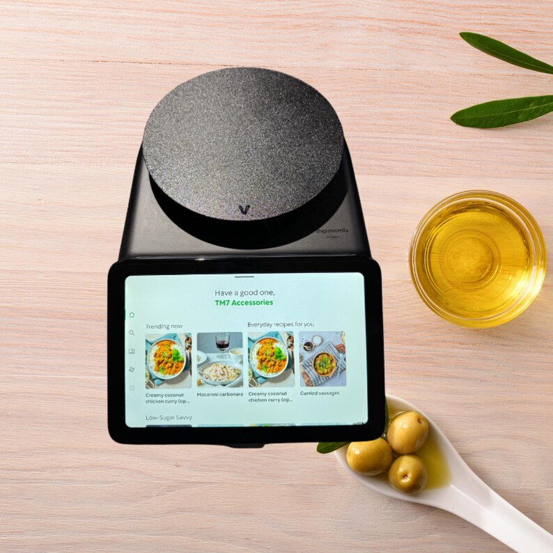 K&ouml;nnte beinhalten: Eine schwarze K&uuml;chenwaage mit digitaler Anzeige, die Rezepte anzeigt. Der Bildschirm zeigt den Text "Have a good one. TM7 Accessories." Eine kleine Sch&uuml;ssel mit &Ouml;l und Oliven befindet sich im Hintergrund.