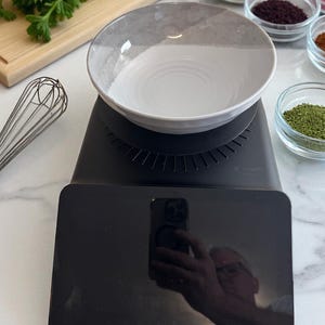 K&ouml;nnte beinhalten: Eine schwarze digitale K&uuml;chenwaage mit einer wei&szlig;-grauen Sch&uuml;ssel. Verschiedene Gew&uuml;rze in kleinen Glasschalen sind rechts angeordnet. Ein Schneebesen und ein Schneidebrett mit Petersilie befinden sich links.