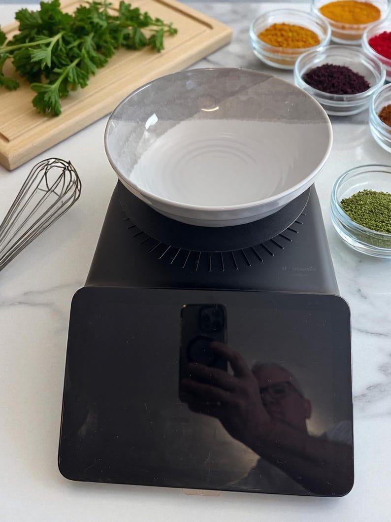 K&ouml;nnte beinhalten: Eine schwarze digitale K&uuml;chenwaage mit einer wei&szlig;-grauen Sch&uuml;ssel. Verschiedene Gew&uuml;rze in kleinen Glasschalen sind rechts angeordnet. Ein Schneebesen und ein Schneidebrett mit Petersilie befinden sich links.