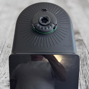 K&ouml;nnte beinhalten: Ein schwarzes K&uuml;chenger&auml;t mit abgerundeter Oberseite und rechteckiger Basis. Die Oberseite hat eine kreisf&ouml;rmige Komponente mit gr&uuml;nem Akzent und dem Wort "thermomix". Es steht auf einer hellen Holzmaserungsoberfl&auml;che.