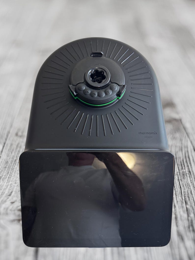 K&ouml;nnte beinhalten: Ein schwarzes K&uuml;chenger&auml;t mit abgerundeter Oberseite und rechteckiger Basis. Die Oberseite hat eine kreisf&ouml;rmige Komponente mit gr&uuml;nem Akzent und dem Wort "thermomix". Es steht auf einer hellen Holzmaserungsoberfl&auml;che.