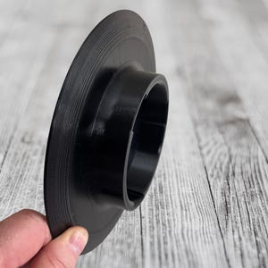 K&ouml;nnte beinhalten: Schwarzer Schallplattenadapter mit einem ausgestellten, zylindrischen Design. Der Adapter hat einen gerillten Au&szlig;enrand und eine zentrale &Ouml;ffnung. Das Objekt wird in der Hand einer Person vor einem hellen Holzmaserungshintergrund gehalten.