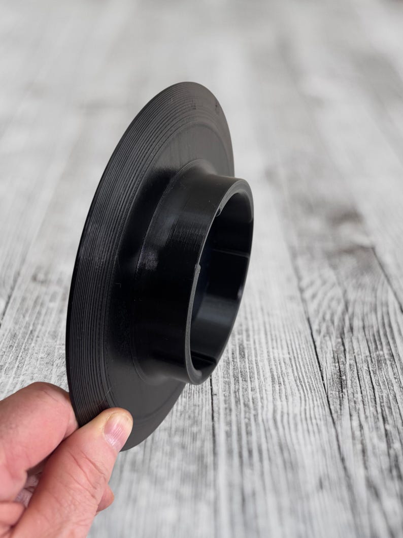K&ouml;nnte beinhalten: Schwarzer Schallplattenadapter mit einem ausgestellten, zylindrischen Design. Der Adapter hat einen gerillten Au&szlig;enrand und eine zentrale &Ouml;ffnung. Das Objekt wird in der Hand einer Person vor einem hellen Holzmaserungshintergrund gehalten.