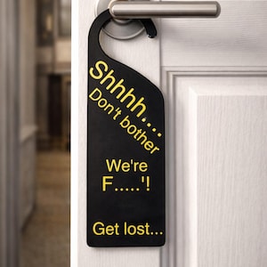 May include: Black door hanger with yellow text that reads "Shhhh... Don't bother We're F.....! Get lost...". The door hanger is hanging on a white door with a silver door handle.