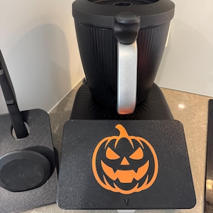 May include: Black coffee maker with a silver handle and a black base. A black rectangular plate with an orange jack-o'-lantern design is in front of the coffee maker. A black utensil holder is to the left.