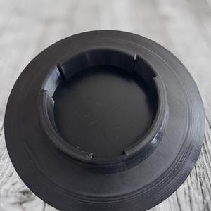 K&ouml;nnte beinhalten: Ein schwarzes, kreisf&ouml;rmiges Objekt mit geriffeltem Au&szlig;enrand und vertieftem Zentrum. Das Objekt hat eine Reihe von gleichm&auml;&szlig;ig verteilten, konzentrischen Rillen. Die Mitte hat vier gleichm&auml;&szlig;ig verteilte Aussparungen.