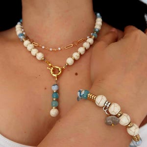 Può includere: Un set composto da collana e bracciale a strati con perline bianche marmorizzate, perline azzurre traslucide e dettagli in metallo dorato. La collana presenta una chiusura a T centrale dorata con un ciondolo pendente, mentre il bracciale include un piccolo ciondolo in pietra grigia.