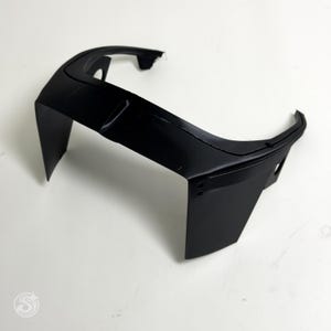 May include: A black, angular automotive part, possibly a spoiler or trim piece, is displayed against a white background. The object has a curved top section and two vertical side panels. The surface appears smooth and matte.
