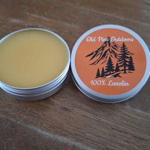Könnte beinhalten: Zwei offene, silberfarbene Dosen mit 100 % Lanolin. Eine Dose ist mit einem hellgelben Balsam gefüllt. Die andere Dose hat einen orangefarbenen Deckel mit dem Text "Old Pine Outdoors" und einer Berg- und Baumgrafik.