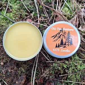 May include: Two open, silver tins of balm on moss and leaves. One tin contains a light yellow balm. The other tin has an orange label with the text "Old Pine Outdoors" and "100% Lanolin" with a mountain and pine tree graphic.