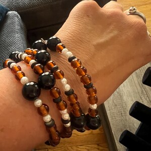 May include: A set of four bracelets with brown, black, and white beads. The bracelets are stacked on a person's wrist.
