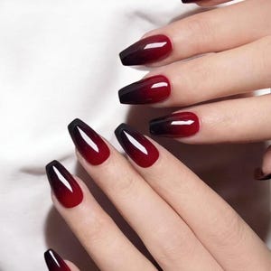 Peut inclure: Gros plan sur une main avec de longs ongles en forme de cercueil. Les ongles présentent un dégradé, passant du noir à la pointe au rouge profond à la base. Les ongles sont brillants et réfléchissent la lumière.