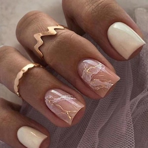 Peut inclure: Ongles carrés avec une base rose clair et un motif marbré doré. Un ongle est peint en blanc uni.