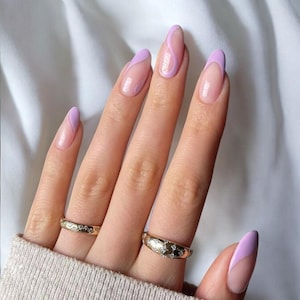 Peut inclure: Mains présentant des ongles en amande avec un bout français violet clair et un motif ondulé. Deux bagues en or avec de petits pierres précieuses sont visibles sur les doigts. Les ongles ont une finition brillante.