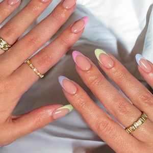 Peut inclure: Gros plan sur des mains avec des ongles en amande arborant une manucure française aux couleurs pastel. Les ongles ont une base nude avec des pointes jaunes, oranges, roses, vertes et bleues. Des bagues dorées ornent les doigts.