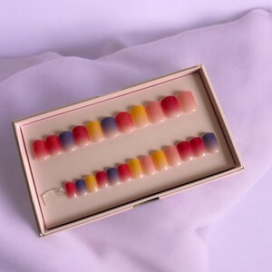 Peut inclure: Un ensemble de 20 faux ongles dans une boîte rose. Les ongles sont courts et de forme carrée. Ils sont ombrés avec un dégradé de jaune, rouge et bleu.