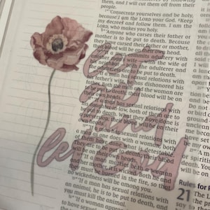 May include: A floral sticker featuring a watercolor-style poppy with a stem and the word "beloved" in a cursive font. The sticker is translucent and placed over a book page.