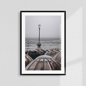 Könnte beinhalten: Ein Schwarzweißfoto einer Straßenlaterne mit einer Lichterkette, die über einer Reihe von durchsichtigen, kuppelförmigen Strukturen hängt. Die Strukturen befinden sich auf einem Pier mit Blick auf das Meer.