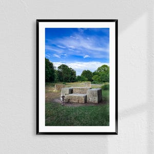May include: Framed photograph of a park scene with a blue sky and scattered clouds. A dog stands near a stone structure in a grassy area. Trees line the background.