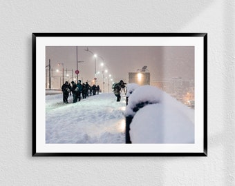 May include: A group of people walking on a snowy street at night. The street is lit by streetlights and there is a snowstorm in the background. The people are wearing winter clothing and some are carrying cameras.