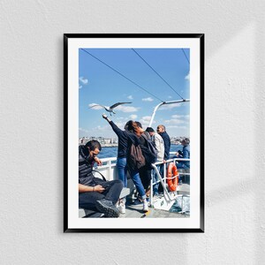 May include: A group of people on a ferry boat, one person is reaching out to feed a seagull flying overhead. The ferry is white with a red life ring. The sky is blue with white clouds.