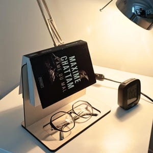 Peut inclure: Un présentoir à livre en bois avec un livre intitulé "MAXIME CHATTAM L'AME DU MAL" et une paire de lunettes. Le livre est posé sur le présentoir, et les lunettes reposent sur la base. Une horloge numérique est visible.