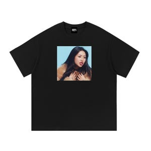 May include: A black short-sleeved t-shirt featuring a square graphic print on the chest. The print displays a person with long dark hair wearing a tan fur-textured garment against a light blue background. The inner neck label reads WRYX.